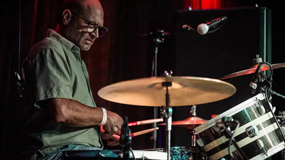 TCC Faculty, Dr. Ted Ward sits at a drum set on a dark stage.