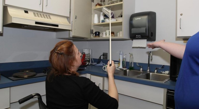 TCC Occupational Therapy Assistant student, seated in a wheelchair, is using a reach and grab tool to retrieve a cup from the cabinet above.